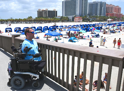 Kyle parks his wheelchair in front of a wooden railing, locating on the left side of the pier. He's wearing a light blue shirt and sunglasses. He look out over the beach, which is lined with people and umbrellas, many of which are blue.