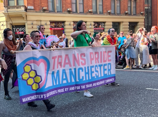 A group of people are walking in Manchester Pride parade, holding a large banner that says, 'Trans Pride Manchester' in pale pink and pale blue lettering.