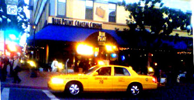 This is a photo of a yellow taxi with the words coffee cab written on the door.  The cab number is 70.  In the background can been the Blue Point Coastal Cuisine Restaurant. 