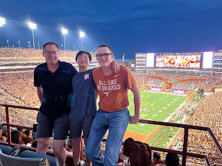 Ken, Tian, and TJ lean on the railings. A packed football stadium, lights, field, and jumbotron behind them.
