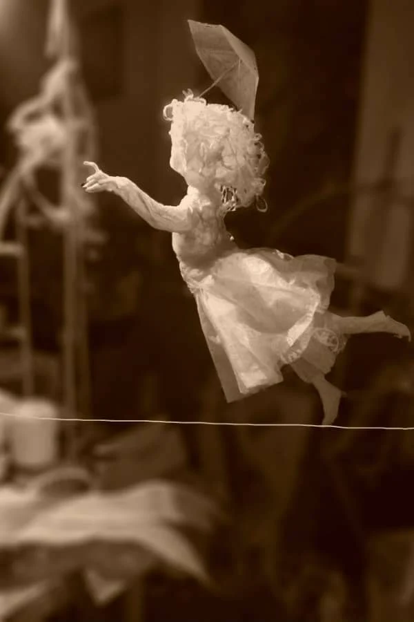 white paper sculpture of young girl walking on tightrope with one arm outstretched and the other holding a parasol
