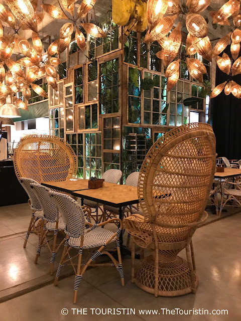 Wooden tables and large hand-woven rattan chairs and black and white bistro chairs in a restaurant with large potted green plants, under a light grey concrete ceiling with about twenty illuminated basket lamps.
