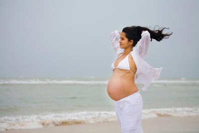 A beautiful, pregnant woman stands on the beach in contemplation as the sea breeze blows her hair in a concept stock photo.