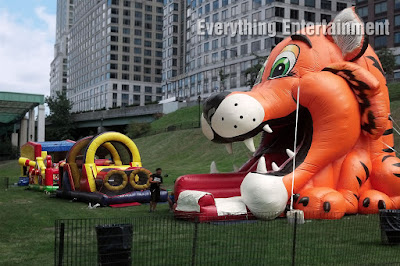 A giant inflatable tiger head slide, obstacle course, and bouncer setup on grass with buildings in the background