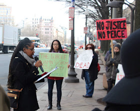 image of demonstration outside Department of Justice office. A woman wearing a coat and scarf with long braids, brown skin, and glasses holds a mic and a clip board in front of 3 people holding protest signs.