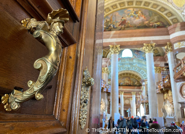 A golden door knob in the form of an angel on a large wooden door leads to a library full of tall wooden bookcases, white marble pillars, and an elaborately painted ceiling.