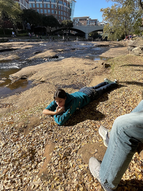 You laying down beside the river in the park in Greensboro, South Carolina on the way to Josh's last soccer game. You are resting your head on your fists. The sun is out and the sky is a deep blue. The river has large boulders scattered throughout it.