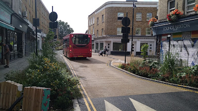 A shopping street with lush planting areas on both sides with a bus driving away in the distance and a person crossing from right to left with a buggy.