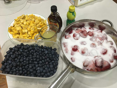 Jam ingredients laid out on a white counter: a plate with a mound of chopped mango, a large glass casserole dish of blueberries, a silver saucepan full of whole strawberries covered with white sugar, a jar of maple syrup and a bottle of lemon juice, and a pyrex measuring cup of lemon juice.