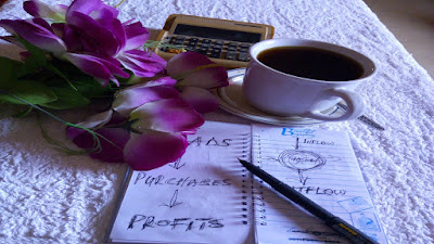 Purple flowers, a calculator, a mug of coffee, a black pen on a note pad on top of a work table.