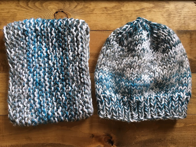 Two white and gray blue variegated knit projects next to each other on a wooden surface. Left one is a cowl. Right one is a hat.