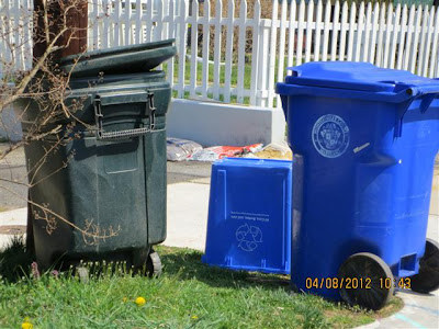 Trash can and recycling containers set on grass next to driveway.