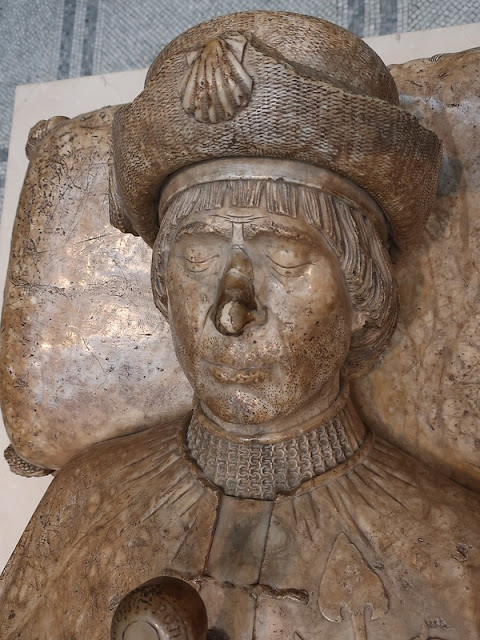 Stone effigy of a man, seen from shoulders up only, head resting on a pillow, hilt of a sword in his hand.