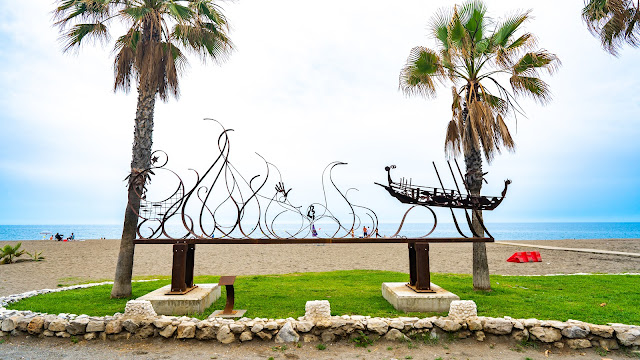 Escultura de hierro en un oasis con palmeras y hierba sobre la arena de la playa con el mar al fondo.