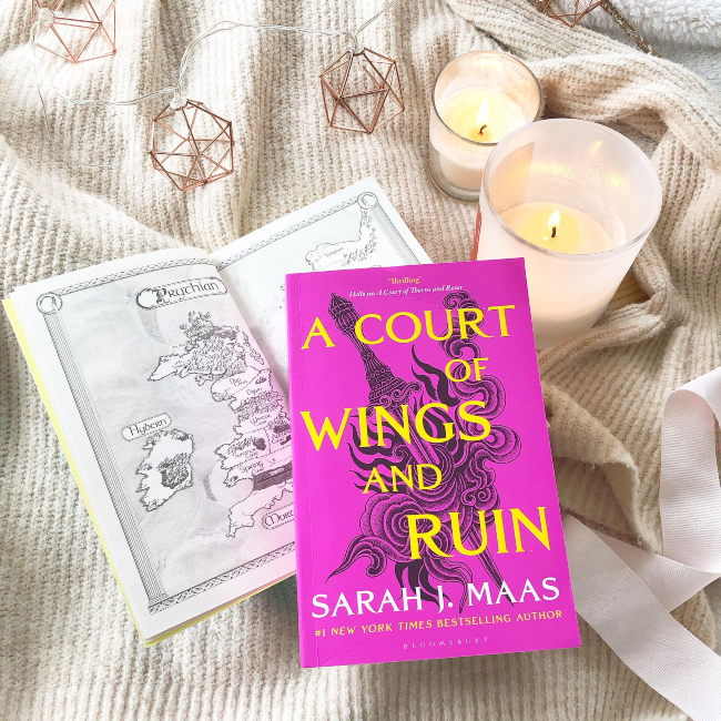 Bright pink cover of 'A Court of Wings and Ruin' by Sarah J Maas on a beige jumper next to a candle