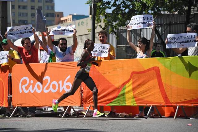 Maratonista queniana Jemima Jelagat Sumgong corre em frente de manifestantes com cartazes de “Fora Temer” no Rio de Janeiro, dia 14 de agosto de 2016.