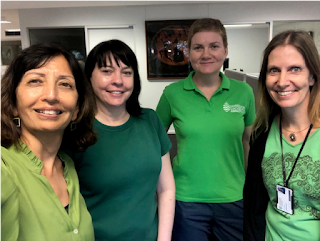 A group selfie image taken in an office space shows four women wearing green clothing. Each person is wearing a different style of green shirt, in a different shade of green. They are all looking up at the camera and smiling