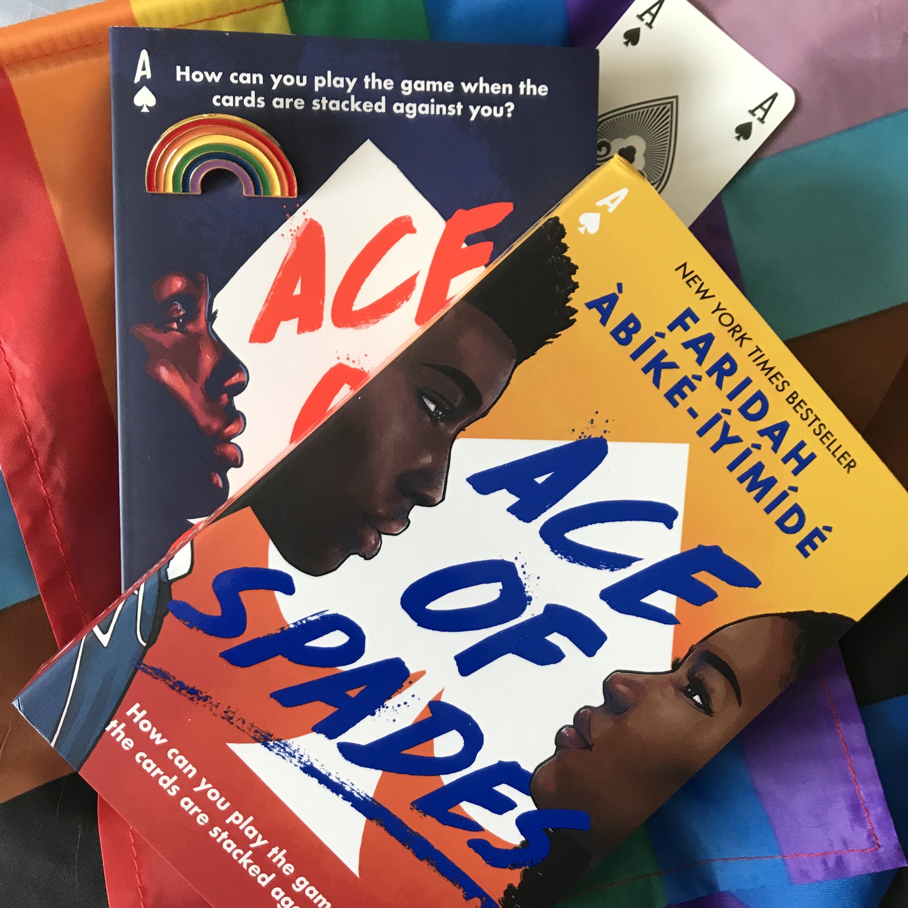 A photo of both editions of Ace of Spades by Faridah Àbíké-Íyímídé; the original edition of the book with the navy blue cover at the bottom, vertical, and the one year anniversary edition with the yellow/orange ombre cover on top, at a diagonal, pointing top right. There is a rainbow pin sittong on the top left corner of the original edition, and a ace of spades playing card poking out from under the two books, top left. They're on a rainbow flag, which is on a larger Pride flag, showing the black and brown stripes.