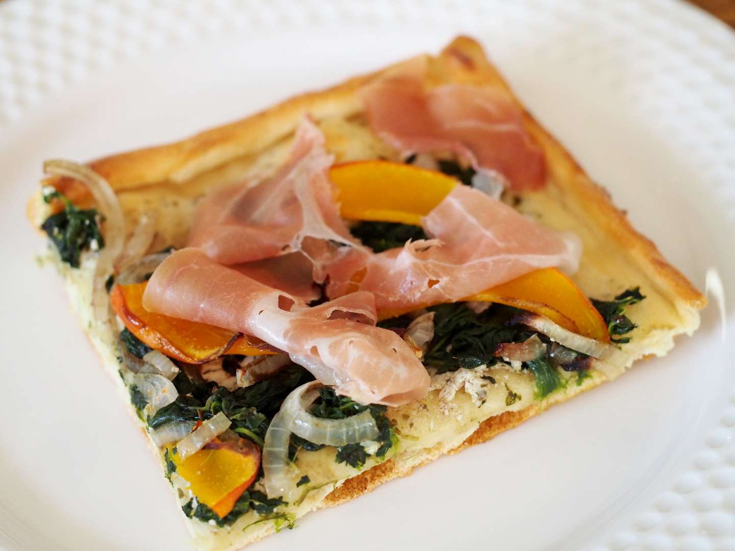 Ein Stück der Kürbispizza auf einem Teller. Der Pizzaboden ist belegt mit Spinat, Zwiebel, Kürbis und rohem Schinken.