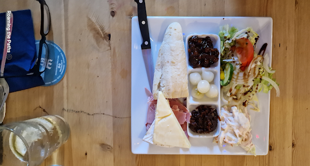 Flatlay of Ploughmans at the Three Mariners with crusty baggette, Mature Cheddar cheese, ham, Brie, pickled onions,  Ploughmans Pickle, Chutney, Colslaw and salad.