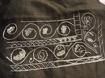 An embroidery sampler with stacked rows of scrolling oak leaves in gold thread on olive-green fabric, showing a variety of fill stitches, and separated by two narrow bands with various small diamond, triangle, and vining fills.