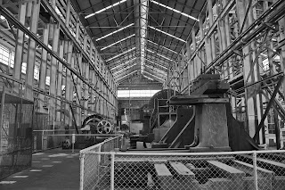 Black and white interior of a large industrial hangar with steel girder frame and fenced-off heavy industrial machinery in foreground and middle distance
