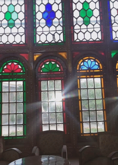A shimmering ray of sunlight passes through colorful stained glass panels set in intricately framed wooden windows, casting vibrant reflections inside a historical Persian building.