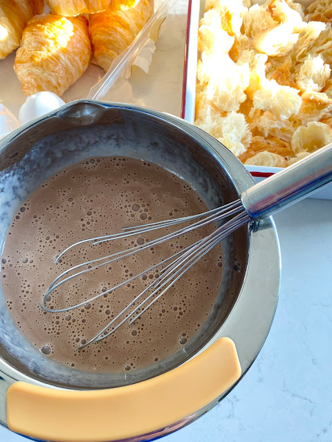 Stainless steel bowl of chocolate milk with croissants in the background.