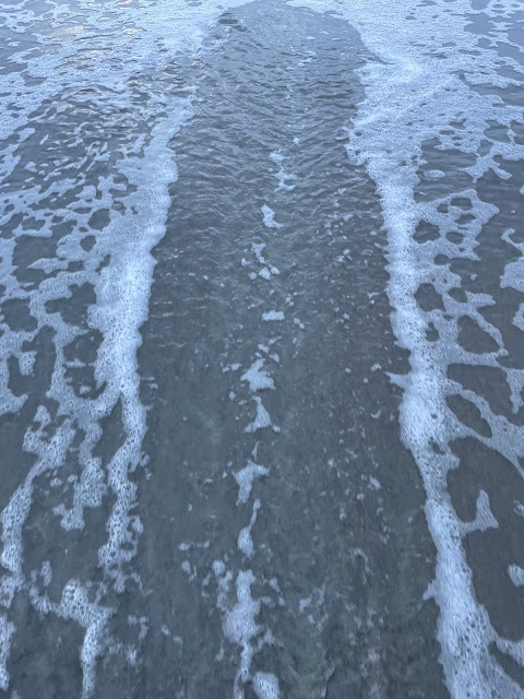 A view of a wave as it moves back to the ocean. A shell or something must have been dragged back with the water because there is a streak of clear water down the middle of the picture with bubbly water all around it. The cleared part is making a path back out to the ocean.