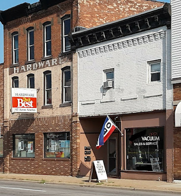 1900s era storefronts. Hardware store beside a vacuum shop. On the sidewalk in front of the vacuum ship is a sign that says, "WE SELL STUFF THAT SUCKS."