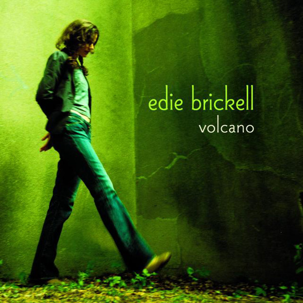 Cover to Edie Brickell album 'Volcano' showing her at the corner of two green walls kicking at some leaves on the ground