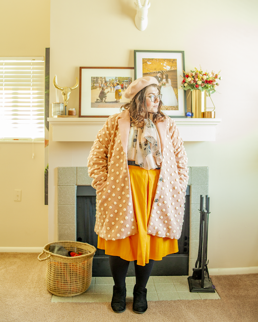 An outfit consisting of a pastel pink beret, a pink knee length pom pom coat over a white sweater tucked into a yellow midi skirt and black ankle boots.