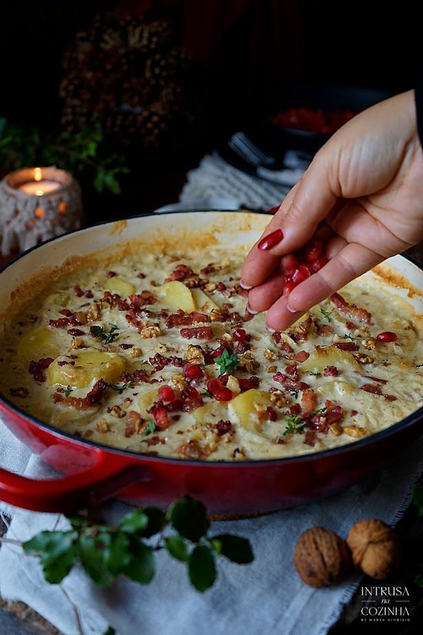 Frigideira de ferro vermelha com batatas gratinadas, e uma mulher a retirar um pouco com uma colher, uma vela com macrame ao fundo e uma coroa de pinhas, ambiente natalicio, darkmood photography, food styling