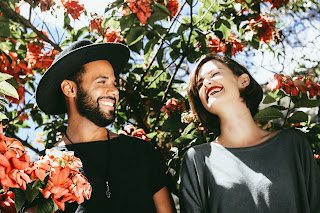 A couple is smiling in the sunshine surrounded by trees full of red blossoms.