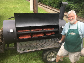 Man with beard wearing shorts, t-shirt, and green apron stands in front of a large barbecue smoker filled with racks of ribs