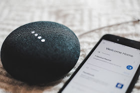 A black, Google Home Mini lies next to a smart phone. The phone's screen reads: "Welcome Home"