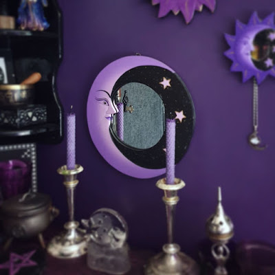 The altar is in the foreground, slightly out of focus; it is on a berry purple altar cloth with a crinkle texture that is on a diagonal to the plane of view. Two silver candle-sticks are upon it, in a classical style, with rolled beeswax purple candles. On the left of the altar is a black plaque displaying a purple pentagram, a small cast iron cauldron with a pentacle on the front, and a partially visible purple glass goblet, cropped out of frame. In the centre is a clear glass candle-holder with a crescent man-in-the-moon and star design moulded into the glass. On the left is an incense censer in the shape of a stylised goddess. Behind the altar and above it is the round crescent moon mirror, it is quite large with a purple crescent moon occupying most of the left of the frame, the right being black with three small stars; the mirrored glass itself is round. The mirror is in focus. To the left of the image the black corner shelving unit is partially visible; the bottom tier has a black stone box with celtic knotwork, a brass Nepalese singing bowl with wooden striker, and partially visible fine-liner print; the print is cropped at the edge of the photograph so the image of the print is not apparent. The second tier is partially visible, with the bottom of the figurine of a wizard in blue, gold and purple robes visible. The shelving unit is slightly out of focus, but the items are identifiable. On the right of the image, a small round moon mirror with a frame consisting of a blueish purple crescent moon and a halo of purple rays is visible, as is a filigree moon pendant with synthetic round stone hanging from it. At the top of the image the halo of rays from the bright purple medium mirror is partially visible. Only the large crescent moon mirror in the centre of the image is in focus, and deliberately so. The photograph is taken in daylight and the shine on metallic objects is quite visible; the black wall is visible in the central mirror, but the wall looks grey in bright direct sunlight. 