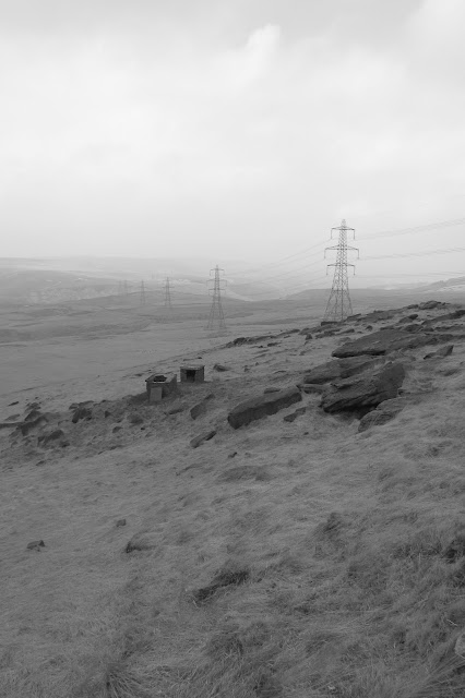 A chain of pylons disappearing into the hazy distance. Wicken Low is barely visible in the distance but gristone and ruined industrial buildings are in the foreground. (B&W)