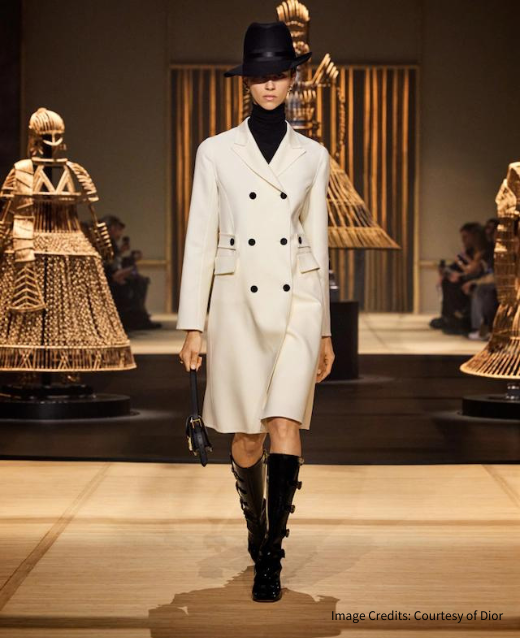 Photo of a female model walking the runway for Dior at Paris Fashion week. Model is wearing a black turtle neck top underneath a cream trench coat with black buttons buttoned up, a black trilby hat and black knee high boots with buckles.