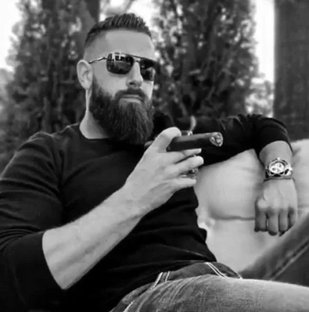 Black and white photo of very handsome man when a long sleeve dark shirt smoking a cigar with sunglasses on outside