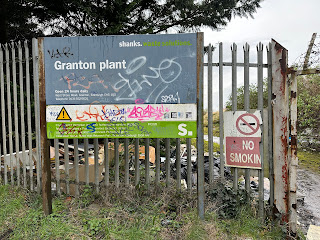 View of a metal fence with two signs on it.  One reads Shanks Waste Solutions, Granton Plant, and the other is a No Smoking sign that is broken in half. Photo by Kevin Nosferatu for the Skulferatu Project.