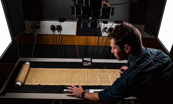 Photograph of man with back to camera in black shirt looking over long yellowed scroll in front of machinery with many cables.