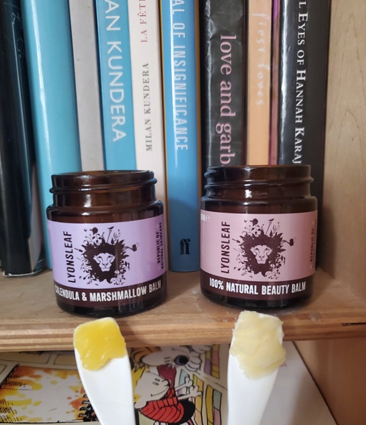 Lyonsleaf Beauty and Calendula Marshmallow balms on spatulas side by side on a bookshelf with plastic free jars showing off pastel packaging