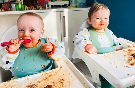 twins sitting in ikea antiplop highchairs with coverall bibs and scoop bibs covered in mess. One has a spoon in their mouth