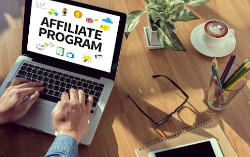 A computer with affiliate program on the screen, notepad, a potted plant, eye glasses, smartphone, a glass pencils holder and a mug of creamy coffee.