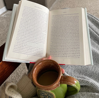 a woman's hand holding a hardcover book open with a cup of coffee, green pottery mug, in her lap