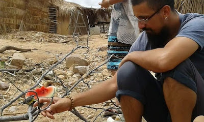 Leandro estende um dedo para um passarinho num galho. Perto, um pedaço de melancia. Um homem próximo, onde só aparece parte da perna. Acredito que seja em uma aldeia.