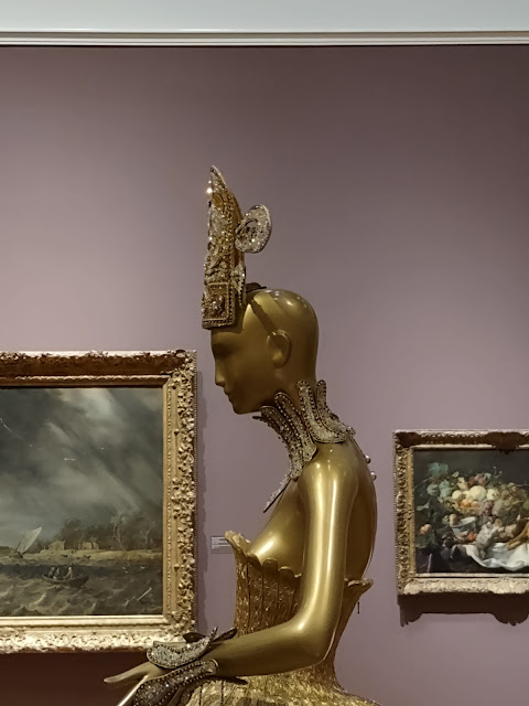 Photo taken in a museum. Mauve wall with two paintings. To the left is a stormy seascape in an elaborate frame, to the right, a still life with fruits and vegetables on a table. In between is a gold mannequin with an elaborate headdress, bodice, and gloves, also in gold.