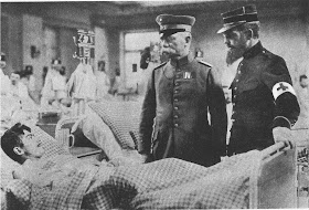 Black and white photo of a military hospital with a man lying on a bed in the very front of the photo. Slightly right to the man are standing two military generals. One on the right is wearing a German military uniform and another one on the right is wearing a French military uniform with a cross symbol on his arm. Behind these three men are nurses and other patients going in and out of the room.