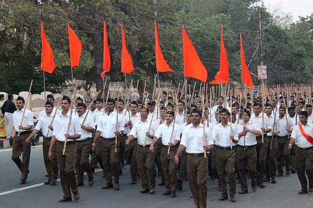 Group of men in matching brown pants and white shirts, carrying sticks and red flags, marching in the street